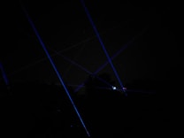 Blue laser beams crisscross against a dark night sky, creating a dramatic and futuristic effect. The beams appear to originate from multiple points, possibly placed on the ground or on structures, and some foliage is faintly visible at the bottom of the image.