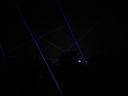 Blue laser beams crisscross against a dark night sky, creating a dramatic and futuristic effect. The beams appear to originate from multiple points, possibly placed on the ground or on structures, and some foliage is faintly visible at the bottom of the image.