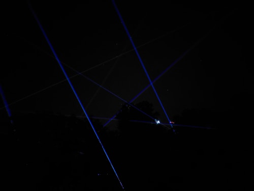 Blue laser beams crisscross against a dark night sky, creating a dramatic and futuristic effect. The beams appear to originate from multiple points, possibly placed on the ground or on structures, and some foliage is faintly visible at the bottom of the image.