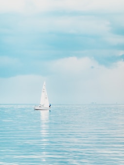 A small sailboat glides across a calm, expansive body of water under a soft, cloudy sky. The horizon is nearly indistinct, blending the sea and sky in gentle hues of blue. The sailboat is centered in the image, with its white sail catching a gentle breeze, and a person can be seen on board.