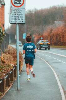 A person wearing sports attire is jogging along a roadside path. A road sign indicates no motor vehicle parking or loading is allowed on the footway or cycle track. In the background, a car is traveling along the adjacent road, and there are trees and bushes along the pathway.