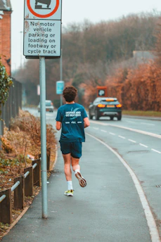 A person wearing sports attire is jogging along a roadside path. A road sign indicates no motor vehicle parking or loading is allowed on the footway or cycle track. In the background, a car is traveling along the adjacent road, and there are trees and bushes along the pathway.