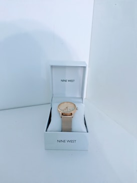 A wristwatch with a gold-tone mesh band and a white dial is displayed inside a white Nine West branded box. The watch is positioned on a cushioned support within the box, set against a simple, plain background.