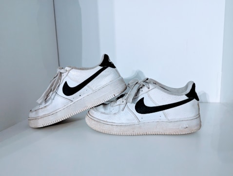 A pair of well-worn, white sneakers with a black swoosh logo displayed on a light gray surface. The shoes have visible signs of wear and dirt, especially on the soles and around the toe area. The laces are loosely tied, and the sneakers are positioned in a casual arrangement.