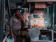 A man stands next to a small street food stall, smoking a cigarette. The stall is equipped with various cooking utensils and ingredients, including a menu displaying prices. A warm light illuminates the scene, casting a cozy glow. There is a helmet hanging on the stall and plastic bags filled with items.