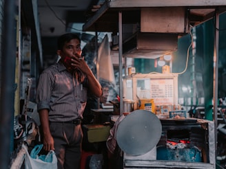 A man stands next to a small street food stall, smoking a cigarette. The stall is equipped with various cooking utensils and ingredients, including a menu displaying prices. A warm light illuminates the scene, casting a cozy glow. There is a helmet hanging on the stall and plastic bags filled with items.