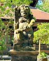 A large, intricately carved stone statue stands in a lush garden setting. The statue depicts a mythical creature with a fierce expression, adorned with ornate patterns and holding a club. Robust trees surround the statue, and a traditional building with a tiled roof is visible in the background.
