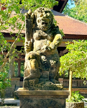 A large, intricately carved stone statue stands in a lush garden setting. The statue depicts a mythical creature with a fierce expression, adorned with ornate patterns and holding a club. Robust trees surround the statue, and a traditional building with a tiled roof is visible in the background.