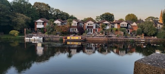 Several suburban houses line a serene waterfront, each with landscaped gardens and a dock extending into the water. Two boats are moored along the shoreline—a white one and a colorful barge. The calm water reflects the houses and surrounding greenery, creating a picturesque and peaceful scene.