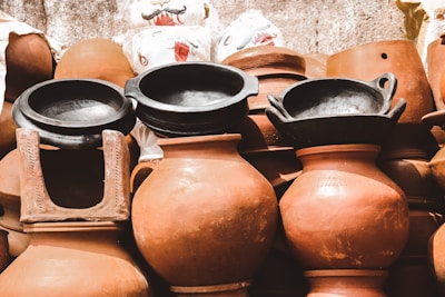 A collection of various clay pots and traditional cookware is displayed. The pots have a reddish-brown hue, while two black clay vessels are prominently placed on top. The background includes sacks, some with decorative elements, adding to the rustic and handcrafted feel.