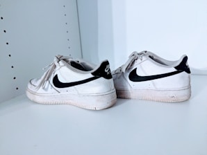 A pair of white athletic shoes with black swoosh logos, placed on a white shelf. The shoes show signs of wear, with visible creases and slight dirt marks.