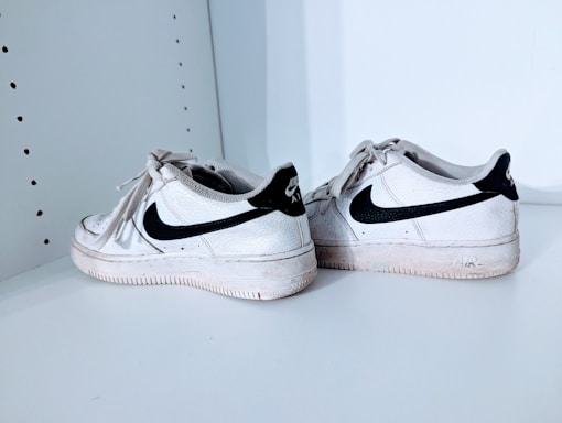 A pair of white athletic shoes with black swoosh logos, placed on a white shelf. The shoes show signs of wear, with visible creases and slight dirt marks.