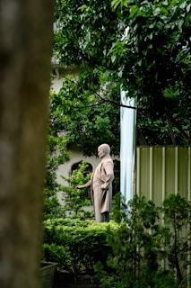 A statue of a historical figure stands amidst lush greenery, surrounded by tall, leafy trees. The statue appears to be made of stone and depicts a man in a suit with his hand extended outward. The background includes a building partially obscured by vegetation.