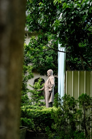 A statue of a historical figure stands amidst lush greenery, surrounded by tall, leafy trees. The statue appears to be made of stone and depicts a man in a suit with his hand extended outward. The background includes a building partially obscured by vegetation.
