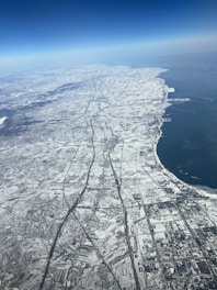 An aerial view showcasing a vast, snow-covered landscape. Geometric agricultural fields and roads are visible, leading towards a coastline with a large body of water on the right. The contrast between the land and water creates a striking visual, while the deep blue sky arcs above.