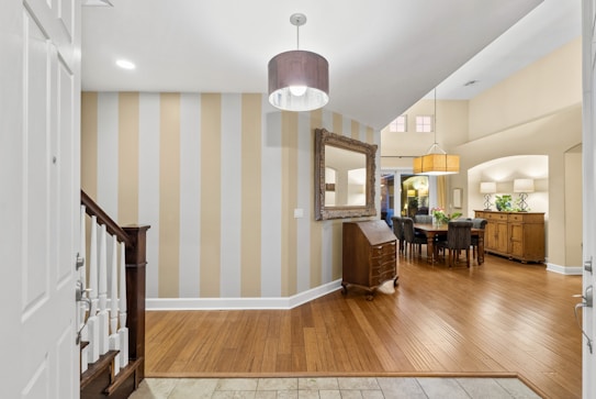 An interior view of a room showcasing a wooden floor with a mix of white and beige vertical striped wall on the left. There is a large mirror with an ornate frame mounted on the wall. Below the mirror, there is a wooden piece of furniture resembling a small dresser or cabinet. A modern, circular light fixture hangs from the ceiling. To the right, there is a dining area with a long dining table surrounded by chairs, illuminated by a large, rectangular hanging light shade. There is also another sideboard or cabinet with decorative plants and lamps on top. The area is well-lit with ceiling lights.