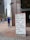 A city street view with a prominent art installation featuring a bold message in large black lettering on a whiteboard. The message reads, 'The only lie I ever told you is that I liked you when I already knew I loved you.' It's placed against a brown stone building, and a few people are walking by in casual attire, including a man in a blue shirt.