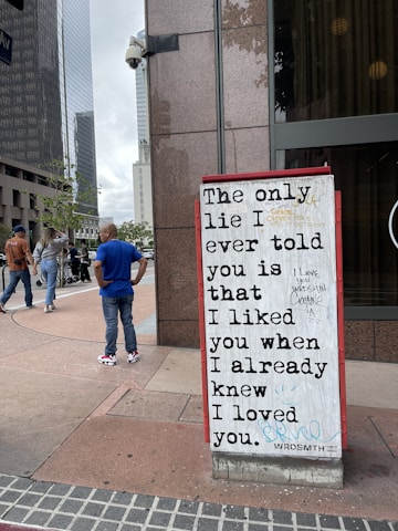 A city street view with a prominent art installation featuring a bold message in large black lettering on a whiteboard. The message reads, 'The only lie I ever told you is that I liked you when I already knew I loved you.' It's placed against a brown stone building, and a few people are walking by in casual attire, including a man in a blue shirt.