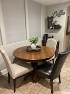 A cozy kitchen corner features a round wooden table with a potted plant centerpiece, surrounded by a beige fabric chair and a black leather chair. The background wall displays a coffee-themed decor with a small shelf holding various items, and a coffee maker is positioned on a console table beneath.