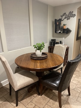 A cozy kitchen corner features a round wooden table with a potted plant centerpiece, surrounded by a beige fabric chair and a black leather chair. The background wall displays a coffee-themed decor with a small shelf holding various items, and a coffee maker is positioned on a console table beneath.