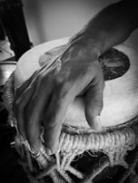 A close-up of a person's hand resting on a traditional drum with a taut rope design. The image is in black and white, emphasizing the texture of both the hand and the drum surface.