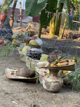 A rural setting with offerings placed on a large tray and a decorated pot. There are various fruits and items like coconuts, bananas, and incense sticks visible. Large green banana leaves and trees form the backdrop, and festive string lights are draped around the setup. A basket filled with more fruits and items sits on the ground.