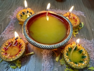 A decorative arrangement of oil lamps, known as diyas, are placed on a wooden surface. Each lamp contains a flickering flame, surrounded by intricate designs and filled with yellow oil. The diyas are adorned with colorful accents, surrounded by scattered flower petals and grains of rice, creating a festive and vibrant atmosphere.
