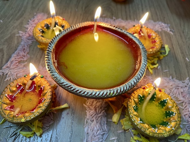 A decorative arrangement of oil lamps, known as diyas, are placed on a wooden surface. Each lamp contains a flickering flame, surrounded by intricate designs and filled with yellow oil. The diyas are adorned with colorful accents, surrounded by scattered flower petals and grains of rice, creating a festive and vibrant atmosphere.