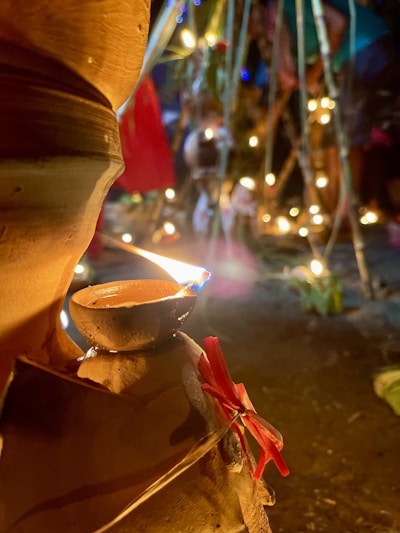 A small clay lamp is lit and placed on a larger clay pot adorned with a red ribbon. The background consists of several glowing lights and blurred figures, suggesting a festive or ceremonial setting. Bamboo sticks and plants are visible, contributing to the decorative atmosphere.