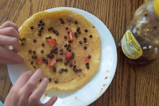 A crepe topped with chocolate chips and sliced strawberries on a white plate. A pair of hands is adjusting the crepe on the plate. A honey bottle is visible on the wooden table to the right.