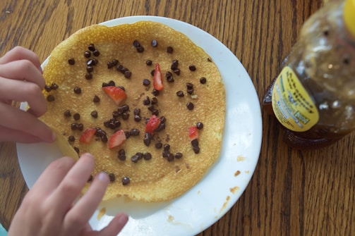 A crepe topped with chocolate chips and sliced strawberries on a white plate. A pair of hands is adjusting the crepe on the plate. A honey bottle is visible on the wooden table to the right.