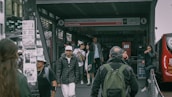 People walking near a TransMilenio bus station entrance with signage indicating the Museo del Oro. Several individuals are wearing jackets and carrying bags. A bus is visible on the right side, and various informational posters are attached to a nearby pole.