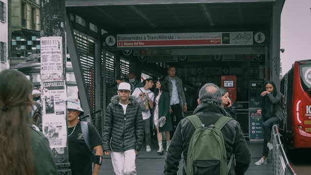 People walking near a TransMilenio bus station entrance with signage indicating the Museo del Oro. Several individuals are wearing jackets and carrying bags. A bus is visible on the right side, and various informational posters are attached to a nearby pole.