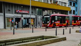 A street scene includes a large red articulated bus labeled 'TransMilenio' parked alongside a building. The building has a storefront with a sign reading 'Supertienda Drogueria Olimpica' and displays metal shutters and glass doors. A person stands outside, possibly using a phone, while a motorcycle is parked nearby. The foreground features a narrow water channel and stone pavement.