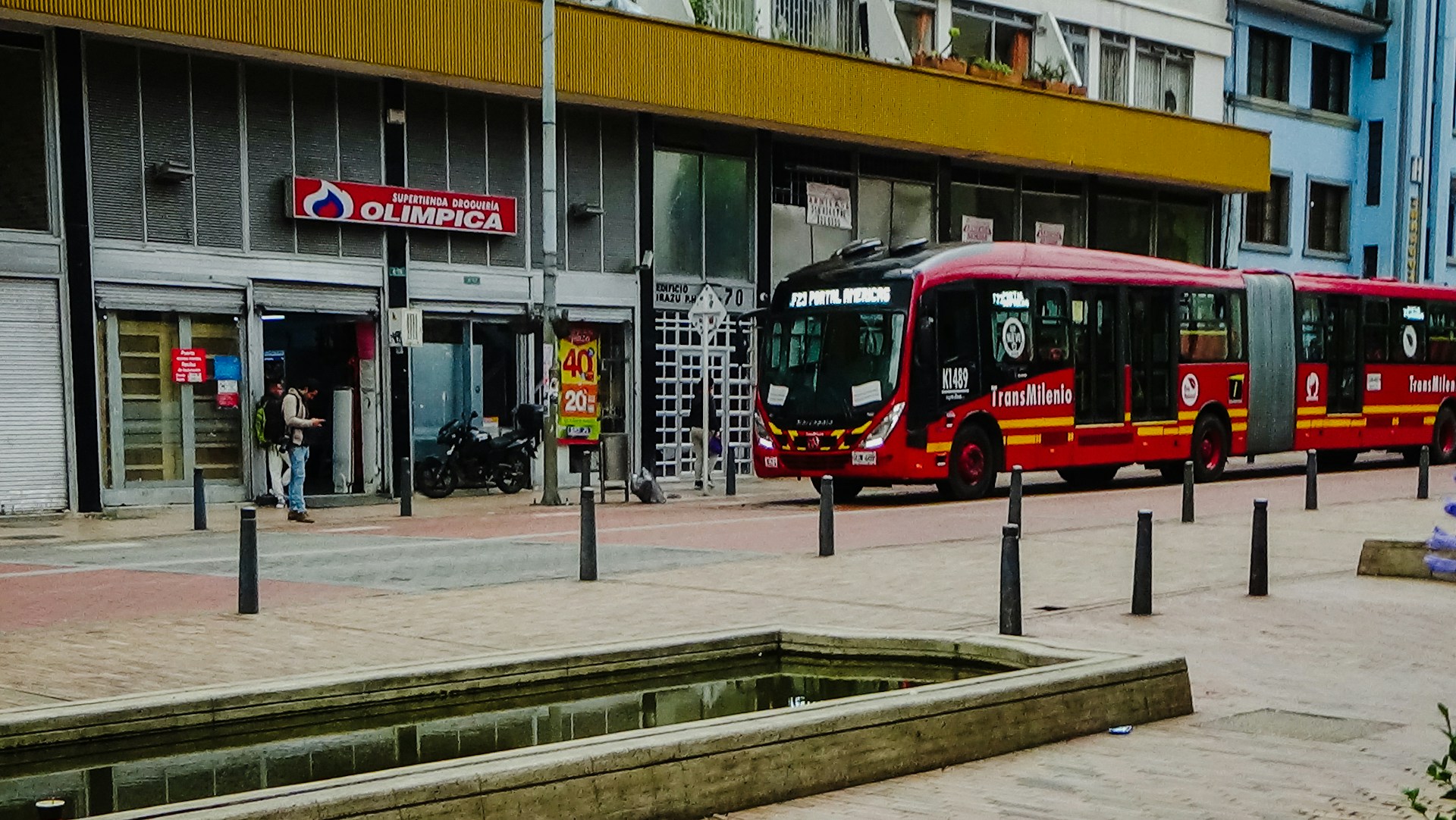 A street scene includes a large red articulated bus labeled 'TransMilenio' parked alongside a building. The building has a storefront with a sign reading 'Supertienda Drogueria Olimpica' and displays metal shutters and glass doors. A person stands outside, possibly using a phone, while a motorcycle is parked nearby. The foreground features a narrow water channel and stone pavement.