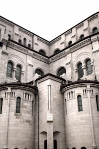 A large stone building with intricate architectural details, featuring multiple arched windows and rounded towers. The structure has a historic and grand appearance, made from light gray stones with dark window frames.