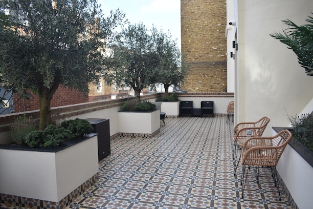 A tiled balcony or terrace area with patterned flooring, surrounded by a tall brick wall and buildings. The space is decorated with several trees in large, square planters and has wicker chairs along the right side. The area appears organized and well-maintained, providing a serene and comfortable outdoor setting.
