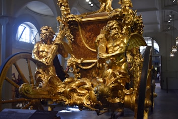 A lavishly decorated, golden carriage adorned with intricate sculptures and mythological figures. The figures are muscular and dynamic, suggesting motion and strength, with detailed carvings of leaves and ornate patterns. The surrounding environment appears to be indoors, likely a museum or gallery with architectural features such as arches and hanging lamps.