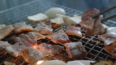 Grilled pieces of meat are sizzling on a wire mesh over an open flame. The meat appears to be pork slices, cooked to a crispy, golden brown. There are also some lighter colored vegetables, likely mushrooms or onions, on the grill. A pair of tongs is holding one piece of meat, showing its texture and color.