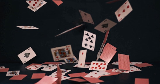 A deck of playing cards scattered mid-air against a dark background, showing various suits and numbers. The cards are dynamically floating, with some already on the surface, creating a sense of motion.