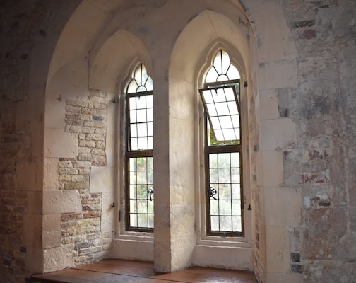 Two arched stone windows with leaded glass panels set into a textured stone wall. The windows have metal detailing and are partially open, allowing natural light to illuminate the stone interior.
