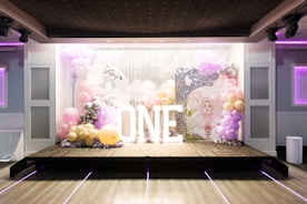A decorative stage setup featuring a backdrop of silver sequins and balloons in pastel colors such as pink, lavender, and gold. Large white illuminated letters spell out 'ONE'. The area is decorated with clusters of balloons and a circular sign with a drawing of a fairy-like character.