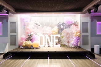 A decorative stage setup featuring a backdrop of silver sequins and balloons in pastel colors such as pink, lavender, and gold. Large white illuminated letters spell out 'ONE'. The area is decorated with clusters of balloons and a circular sign with a drawing of a fairy-like character.