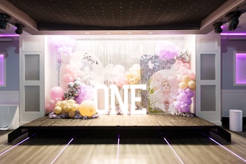 A decorative stage setup featuring a backdrop of silver sequins and balloons in pastel colors such as pink, lavender, and gold. Large white illuminated letters spell out 'ONE'. The area is decorated with clusters of balloons and a circular sign with a drawing of a fairy-like character.