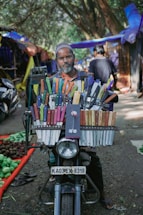 A man is sitting on a motorcycle that is laden with a large display of colorful knives. He is positioned in an outdoor market setting, with stalls and trees in the background. The motorcycle has a visible license plate, and the knives are neatly organized in rows with various colored handles.