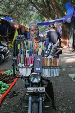 A man is sitting on a motorcycle that is laden with a large display of colorful knives. He is positioned in an outdoor market setting, with stalls and trees in the background. The motorcycle has a visible license plate, and the knives are neatly organized in rows with various colored handles.