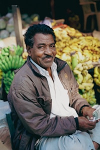 A man wearing a brown jacket and white shirt sits in front of a display of bananas. He appears relaxed and is holding currency notes in his hands. The background includes a variety of fresh produce, primarily bananas, indicating a market setting.