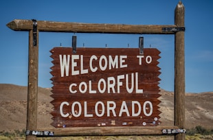 A wooden sign displaying the words 'Welcome to Colorful Colorado' is seen, set against a backdrop of barren hills and a clear blue sky.