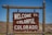 A wooden sign displaying the words 'Welcome to Colorful Colorado' is seen, set against a backdrop of barren hills and a clear blue sky.