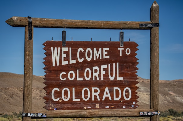 A wooden sign displaying the words 'Welcome to Colorful Colorado' is seen, set against a backdrop of barren hills and a clear blue sky.